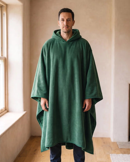 Man wearing the Hazel oversized hooded blanket in forest green, standing in a warm living room, front view