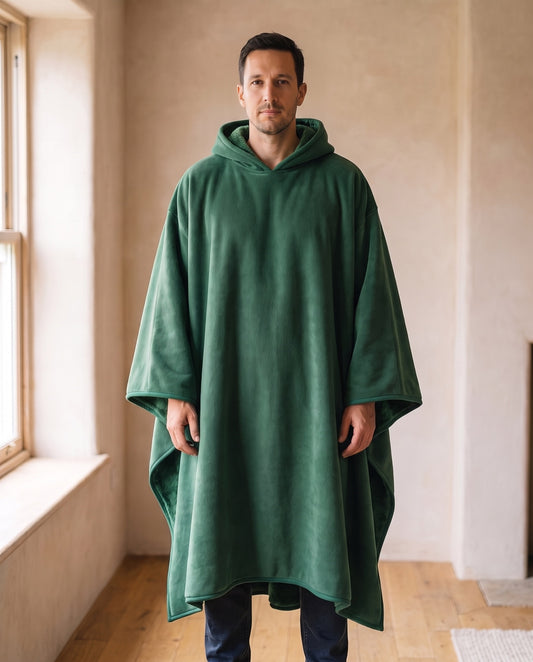 Man wearing the Hazel oversized hooded blanket in forest green, standing in a warm living room, front view