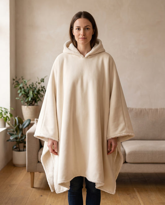 Woman wearing the Wren oversized hooded blanket in soft oatmeal, standing in a warm living room, front view