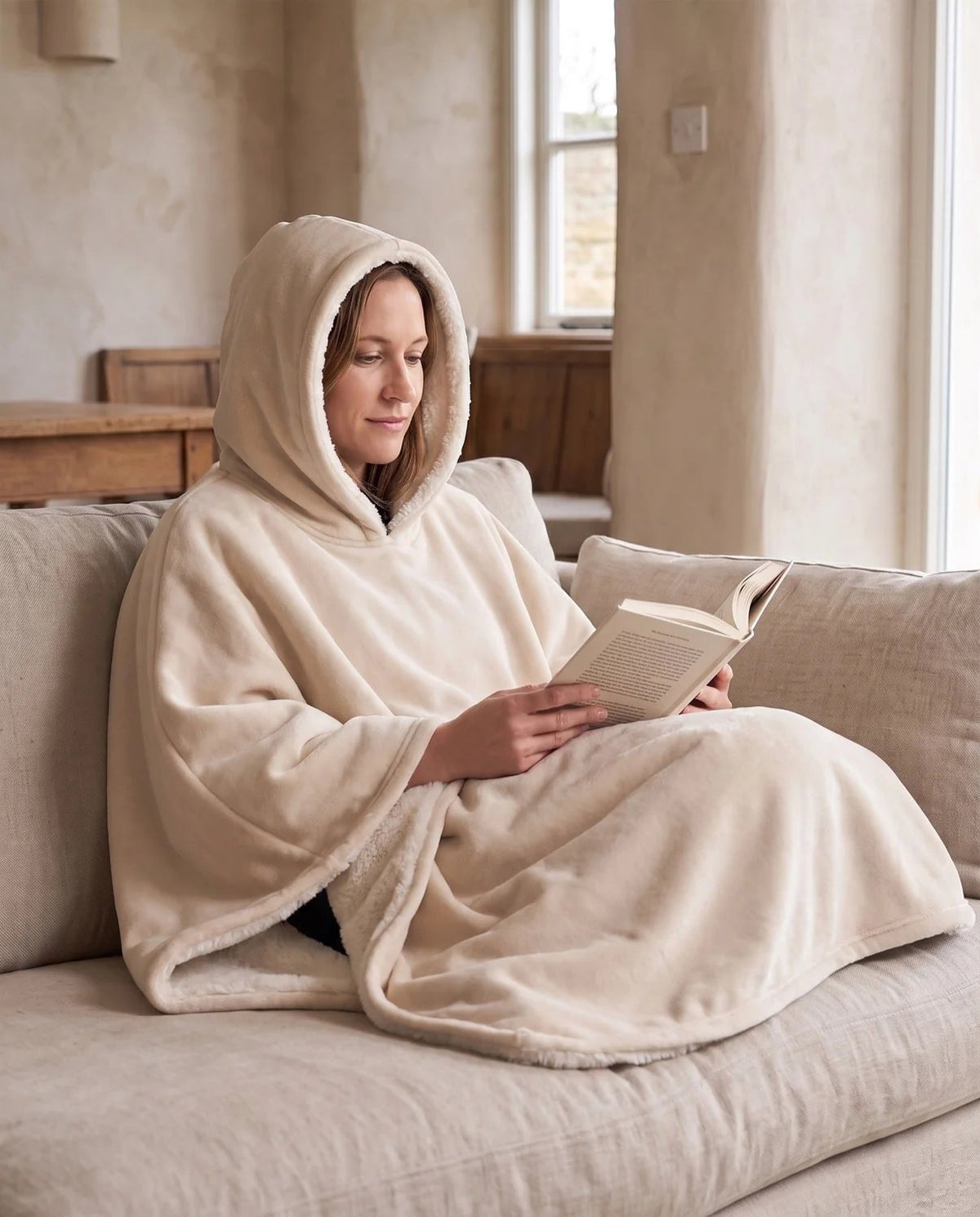 Woman reading on a sofa in lamplight wearing the Wren hooded blanket in oatmeal with the hood up