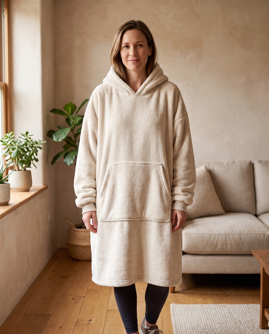 Woman wearing the Wren oversized hooded blanket in oatmeal, standing in a warm living room, front view