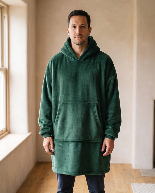 Man wearing the Hazel oversized hooded blanket in forest green, standing in a warm living room, front view