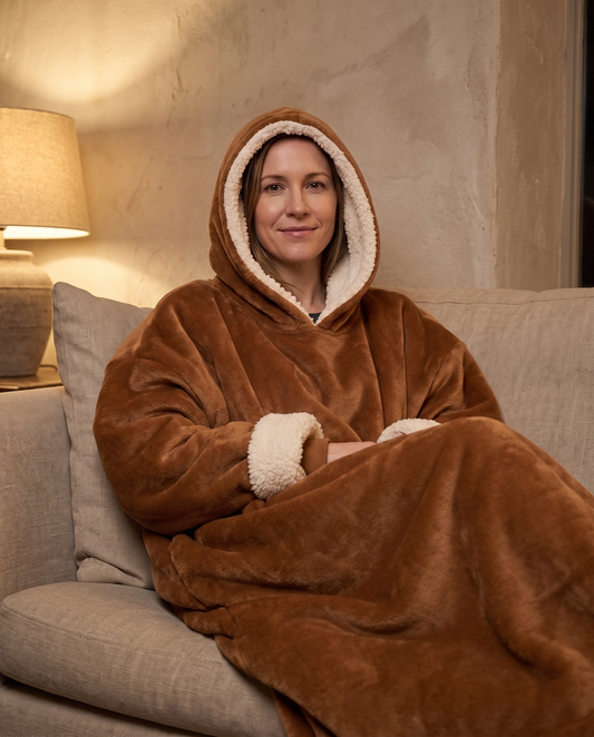 Woman on a sofa in lamplight wearing the Alba hooded blanket in cognac with the sherpa-lined hood up
