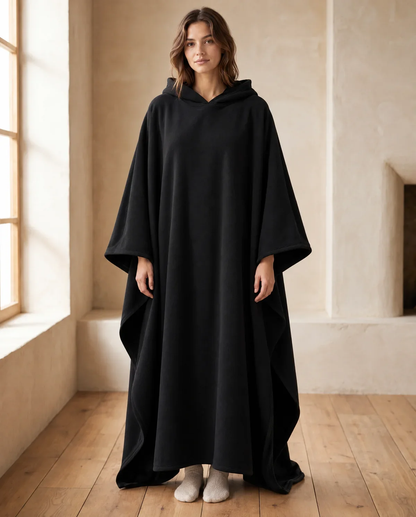 Woman wearing the Juna oversized hooded blanket in black, standing in a warm living room, front view