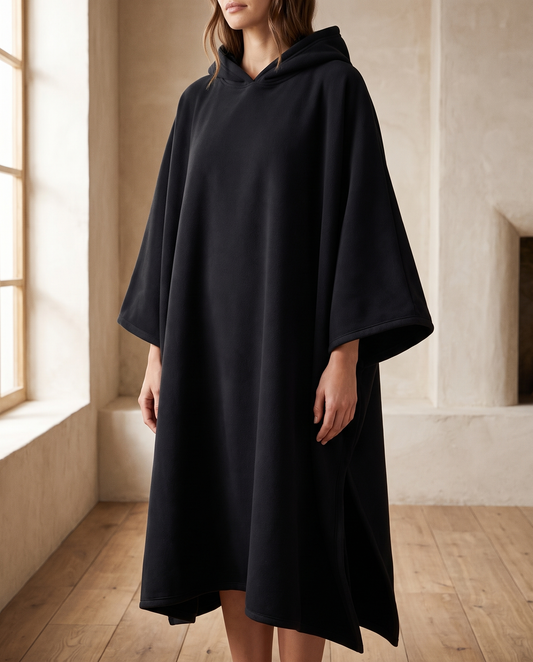 Woman wearing the Juna oversized hooded blanket in black, standing in a warm living room, front view